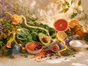 Assorted healthy foods: leafy greens, avocado, citrus, beans, seeds, and salt arranged with a floral backdrop and a glowing light ribbon.