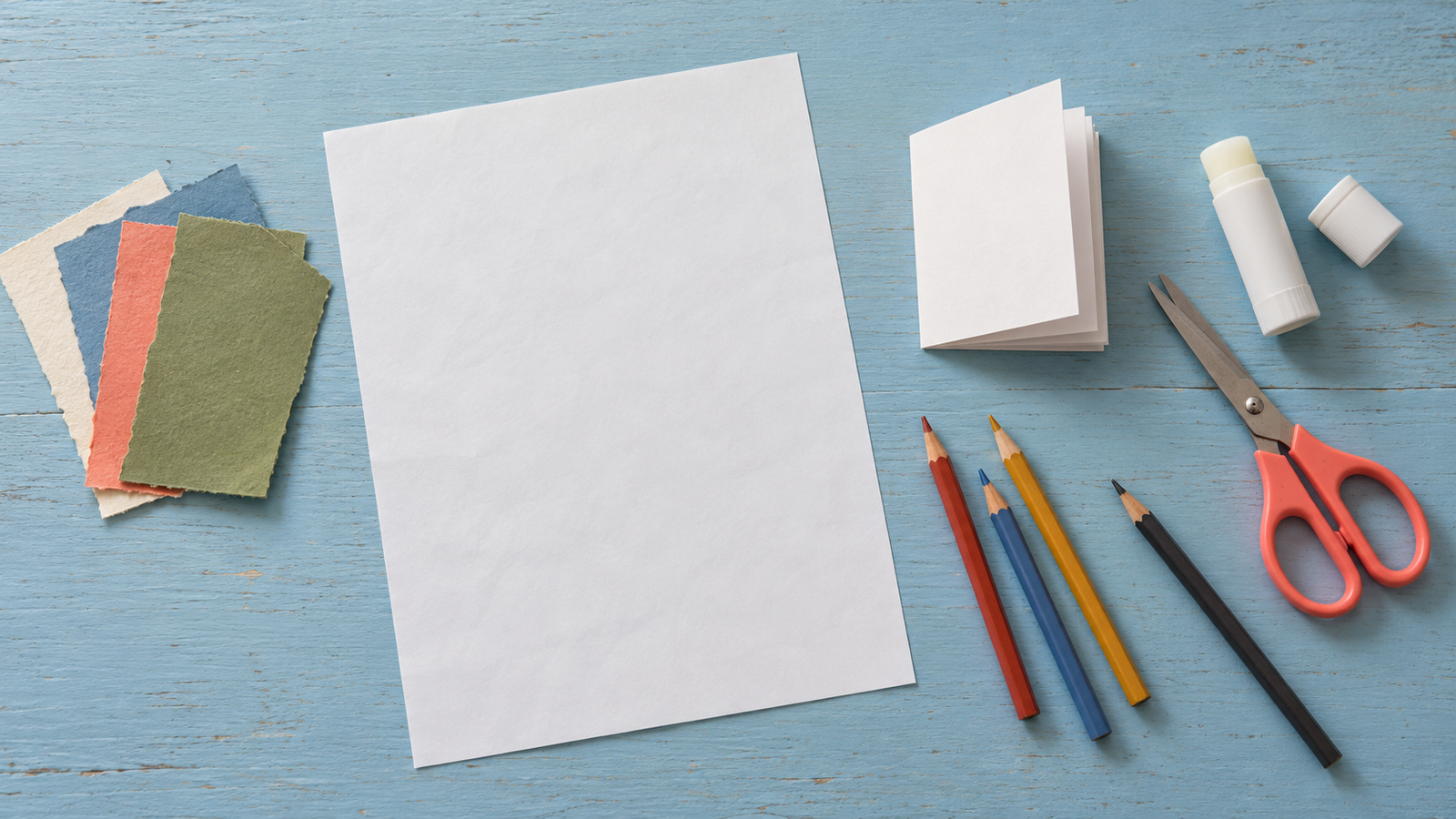 Printer paper, a blank folded booklet, scissors, glue stick, pencils and colored paper scraps on a blue wooden table.