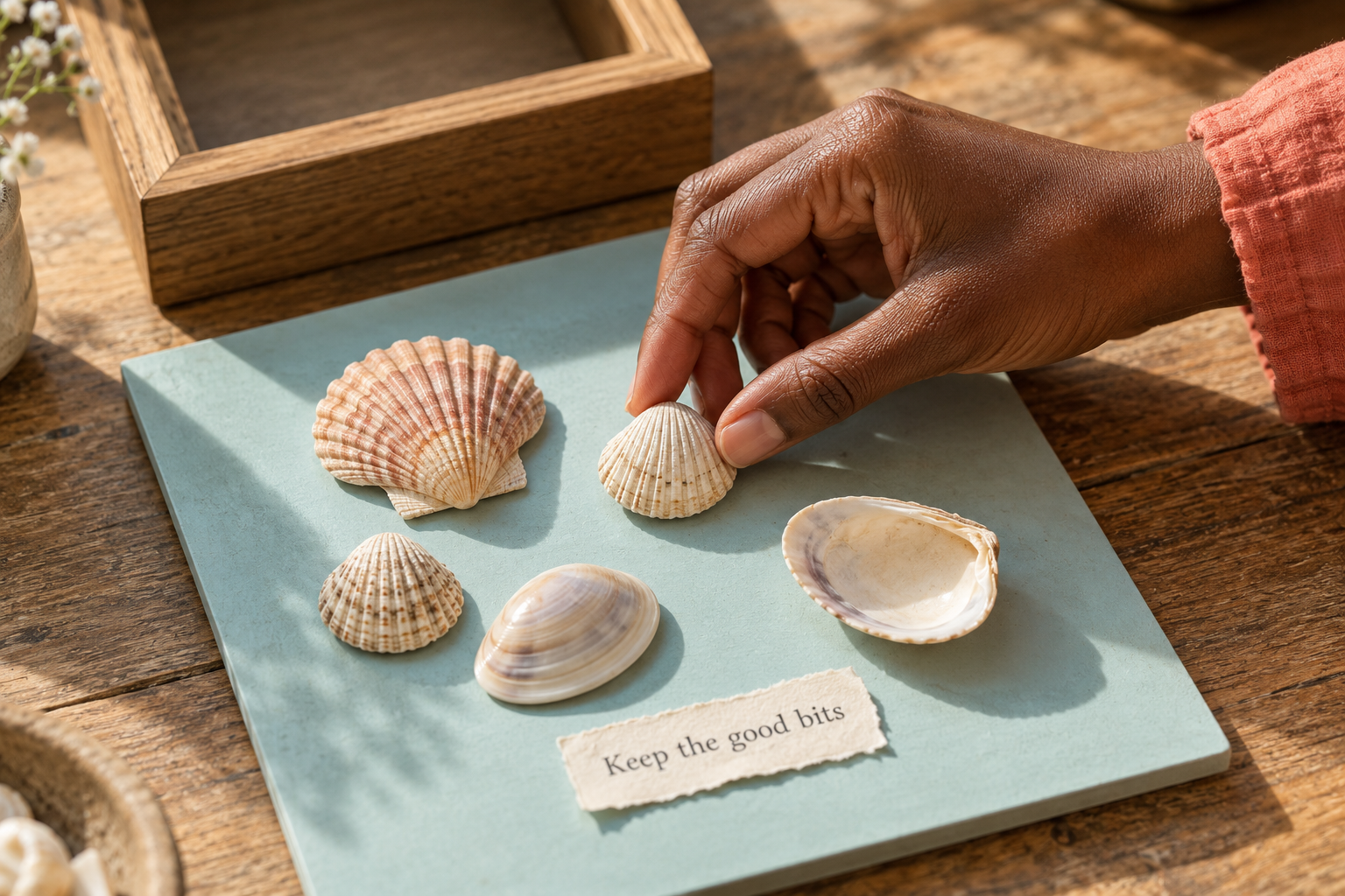 A dark-skinned hand arranging shells on a flat aqua board beside a “Keep the good bits” paper label.